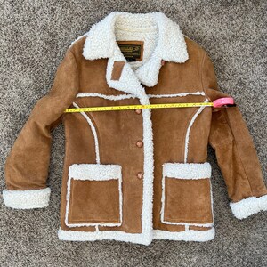 Peut inclure: Une veste en daim marron avec une doublure en sherpa blanche. La veste a une fermeture &agrave; boutons, deux poches et un col. La veste est pos&eacute;e &agrave; plat sur un tapis marron.