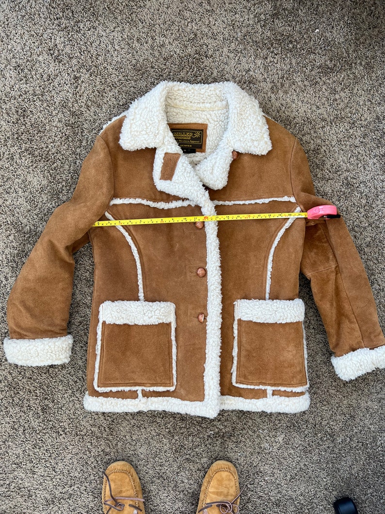 Peut inclure: Une veste en daim marron avec une doublure en sherpa blanche. La veste a une fermeture &agrave; boutons, deux poches et un col. La veste est pos&eacute;e &agrave; plat sur un tapis marron.
