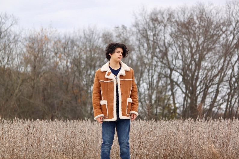 Peut inclure: Une jeune personne portant une veste en daim marron avec une doublure en peau de mouton blanche, un jean bleu et des cheveux fonc&eacute;s. La personne est debout dans un champ d'herbe s&egrave;che avec un arri&egrave;re-plan flou d'arbres.