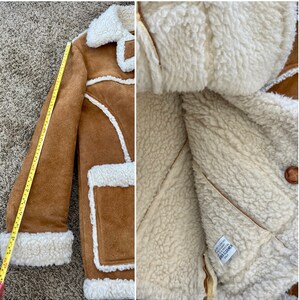 Peut inclure: Une veste en daim marron avec une doublure en peau de mouton blanche. La veste a un col, des manches longues et une fermeture &agrave; boutons. L'&eacute;tiquette &agrave; l'int&eacute;rieur de la veste indique "Cuir v&eacute;ritable, Fabriqu&eacute; en Cor&eacute;e".