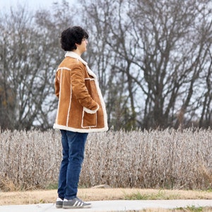 Peut inclure: Une personne portant une veste en daim marron avec une doublure en peau de mouton blanche, un jean bleu et des baskets blanches. La personne est debout devant un champ de plantes s&eacute;ch&eacute;es.