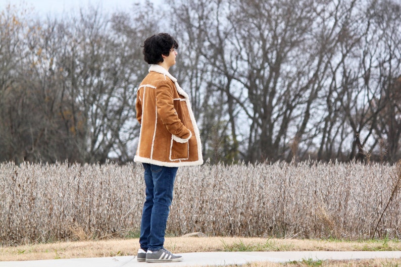 Peut inclure: Une personne portant une veste en daim marron avec une doublure en peau de mouton blanche, un jean bleu et des baskets blanches. La personne est debout devant un champ de plantes s&eacute;ch&eacute;es.