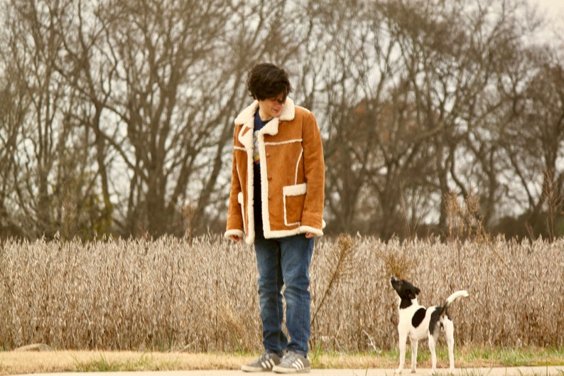 Peut inclure: Une personne portant une veste en daim marron avec un col en fourrure blanche se tient dans un champ d'herbe s&egrave;che avec un petit chien noir et blanc qui la regarde.
