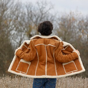 Peut inclure: Une veste en daim marron avec une doublure en peau de mouton blanche. La veste est ouverte et la personne qui la porte se tient dans un champ.