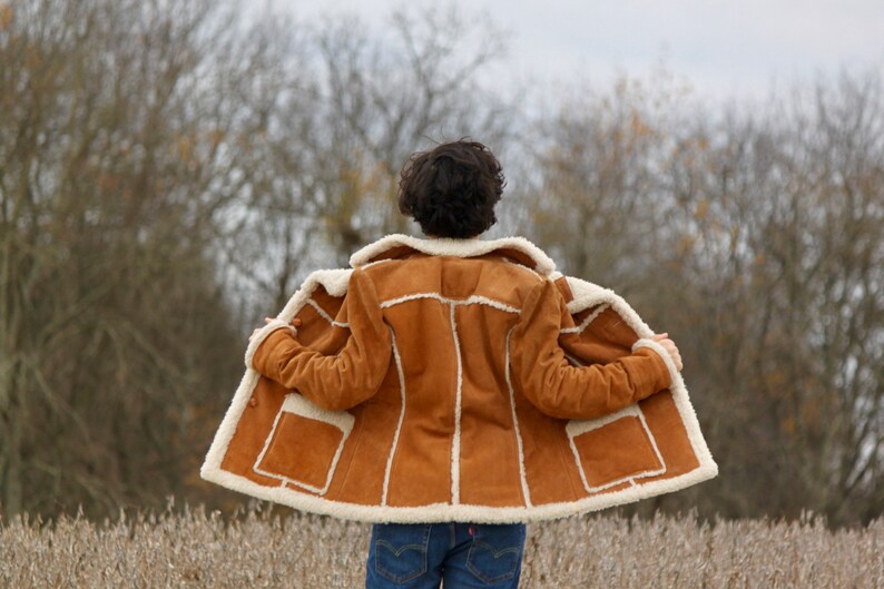 Peut inclure: Une veste en daim marron avec une doublure en peau de mouton blanche. La veste est ouverte et la personne qui la porte se tient dans un champ.