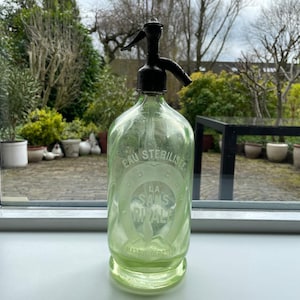 May include: A vintage green glass seltzer bottle with a black spout and top. The bottle has embossed text that reads "EAU STERILISE" and "LA SANS RIVALE". The bottle is sitting on a windowsill.
