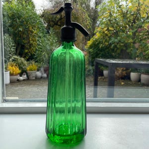 May include: A vintage-style green glass seltzer bottle with a black top and spout. The ribbed glass bottle is cylindrical and sits on a white surface, with a blurred outdoor scene visible through a window in the background.