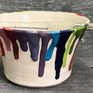 Puede incluir: Un cuenco de cer&aacute;mica blanco con goteos de pintura coloridos en tonos rojos, morados, azules y verdes. Un pincel descansa sobre la parte superior del cuenco. El cuenco tiene un borde festoneado y est&aacute; sobre un fondo oscuro.