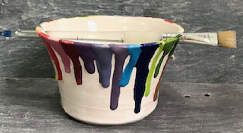 Puede incluir: Un cuenco de cer&aacute;mica blanco con goteos de pintura coloridos en tonos rojos, morados, azules y verdes. Un pincel descansa sobre la parte superior del cuenco. El cuenco tiene un borde festoneado y est&aacute; sobre un fondo oscuro.
