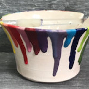 May include: A white ceramic paint holder with colourful paint drips in red, orange, yellow, green, blue, and purple. A paintbrush rests across the top. The holder is approximately 13 cm in diameter and 10 cm tall.