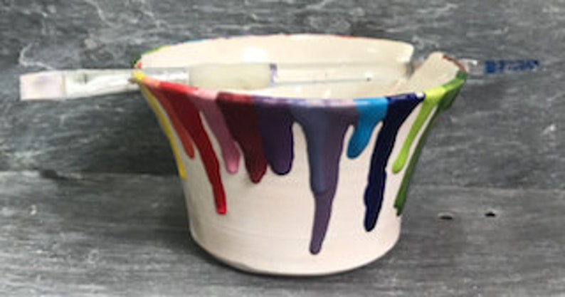 Puede incluir: Un recipiente de cer&aacute;mica blanco para pintura con goteos de pintura coloridos en rojo, naranja, amarillo, verde, azul y morado. Un pincel descansa sobre la parte superior. El recipiente mide unos 13 cm de di&aacute;metro y 10 cm de alto.