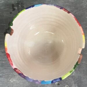 Puede incluir: Un cuenco de cer&aacute;mica hecho a mano con un interior blanco y un borde pintado con un arco&iacute;ris colorido. El cuenco tiene forma redonda y una superficie texturizada, con crestas visibles en el interior. El borde presenta una vibrante gama de colores, incluyendo rojo, naranja, amarillo, verde, azul y morado.