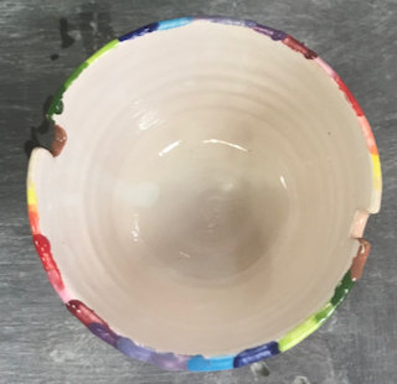 Puede incluir: Un cuenco de cer&aacute;mica hecho a mano con un interior blanco y un borde pintado con un arco&iacute;ris colorido. El cuenco tiene forma redonda y una superficie texturizada, con crestas visibles en el interior. El borde presenta una vibrante gama de colores, incluyendo rojo, naranja, amarillo, verde, azul y morado.