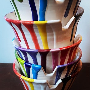 Puede incluir: Una pila de cuatro cuencos de cer&aacute;mica blanca con goteos de pintura colorida en rojo, amarillo, azul, verde, morado y negro. Los cuencos tienen una forma &uacute;nica con un dise&ntilde;o similar a un pico en el lateral. La pintura gotea por los lados de los cuencos.