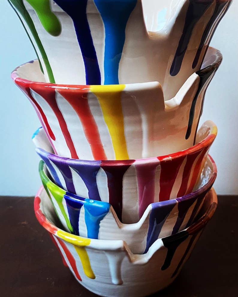 Puede incluir: Una pila de cuatro cuencos de cer&aacute;mica blanca con goteos de pintura colorida en rojo, amarillo, azul, verde, morado y negro. Los cuencos tienen una forma &uacute;nica con un dise&ntilde;o similar a un pico en el lateral. La pintura gotea por los lados de los cuencos.