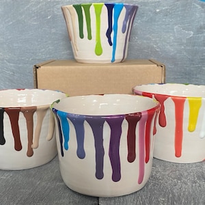May include: A set of white ceramic bowls with colourful paint drip designs. The bowls have drips of various colours, including red, blue, green, and purple, creating a vibrant and artistic look. The bowls are of different sizes.