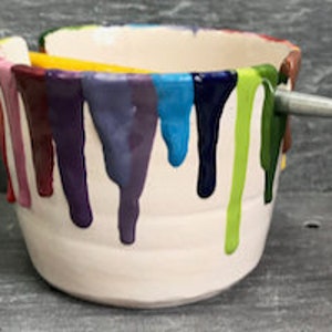 Puede incluir: Un cuenco de pintura de cer&aacute;mica blanca con goteos de pintura coloridos en tonos morado, azul, verde y rosa. Un pincel amarillo y plateado descansa sobre la parte superior del cuenco, que est&aacute; sobre una superficie gris.