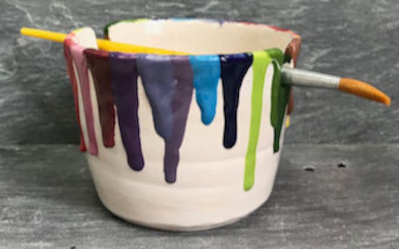 Puede incluir: Un cuenco de pintura de cer&aacute;mica blanca con goteos de pintura coloridos en tonos morado, azul, verde y rosa. Un pincel amarillo y plateado descansa sobre la parte superior del cuenco, que est&aacute; sobre una superficie gris.