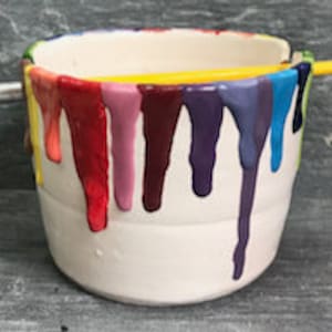 Puede incluir: Un bote de pintura de cer&aacute;mica blanca con goteos de pintura coloridos en rojo, morado, azul y amarillo. Un pincel amarillo descansa sobre la parte superior del bote y otro pincel est&aacute; en el lateral.