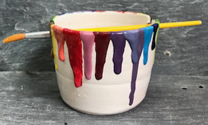 Puede incluir: Un bote de pintura de cer&aacute;mica blanca con goteos de pintura coloridos en rojo, morado, azul y amarillo. Un pincel amarillo descansa sobre la parte superior del bote y otro pincel est&aacute; en el lateral.