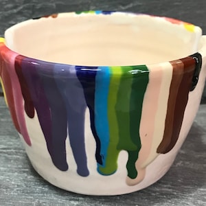 Puede incluir: Un peque&ntilde;o cuenco de cer&aacute;mica blanquecina con un dise&ntilde;o de goteo de arco&iacute;ris. Los colores incluyen rojo, naranja, amarillo, verde, azul y morado. El cuenco tiene un acabado liso y brillante y es adecuado para objetos peque&ntilde;os.