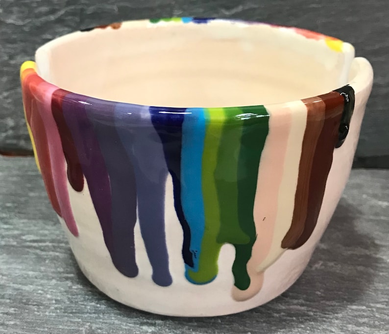 Puede incluir: Un peque&ntilde;o cuenco de cer&aacute;mica blanquecina con un dise&ntilde;o de goteo de arco&iacute;ris. Los colores incluyen rojo, naranja, amarillo, verde, azul y morado. El cuenco tiene un acabado liso y brillante y es adecuado para objetos peque&ntilde;os.