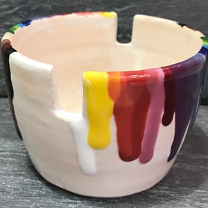 Puede incluir: Un cenicero de cer&aacute;mica con interior y exterior color crema. El borde est&aacute; decorado con un arco&iacute;ris de colores, y goteos de pintura colorida bajan por los lados. El cenicero tiene tres muescas para apoyar los cigarrillos.