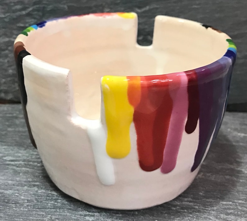 Puede incluir: Un cenicero de cer&aacute;mica con interior y exterior color crema. El borde est&aacute; decorado con un arco&iacute;ris de colores, y goteos de pintura colorida bajan por los lados. El cenicero tiene tres muescas para apoyar los cigarrillos.