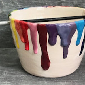 Puede incluir: Un cuenco de cer&aacute;mica blanco con goteos de pintura coloridos en rojo, naranja, amarillo, verde, azul y morado. Un pincel negro descansa sobre el borde superior.
