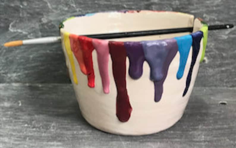 Puede incluir: Un cuenco de cer&aacute;mica blanco con goteos de pintura coloridos en rojo, naranja, amarillo, verde, azul y morado. Un pincel negro descansa sobre el borde superior.