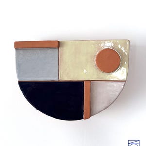 May include: A ceramic wall sculpture with a geometric design. The sculpture is made up of different colored tiles, including gray, black, beige, and orange. The sculpture is in the shape of a half-circle.