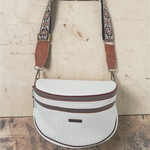 Peut inclure: Sac bandoulière blanc avec un motif texturé, une fermeture éclair marron et une sangle décorative à motif floral. Le sac a une forme incurvée et un petit détail métallique rectangulaire.