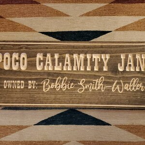 May include: A wooden sign with the text "POCO CALAMITY JANE" and "OWNED BY: Bobbie Smith-Walker" in a light brown color. The sign has a dark brown wood grain and a light brown frame.