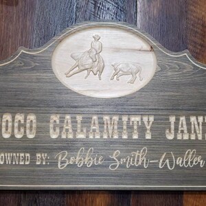 May include: A rustic wooden sign with a carved image of a cowboy riding a horse and a steer. The sign reads "Poco Calamity Jane Owned By: Bobbie Smith-Walker".