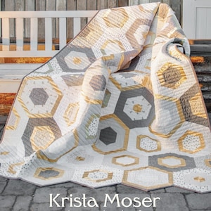 Puede incluir: Una colcha con un patrón de panal de abeja en tonos grises, dorados y blancos. La colcha está doblada en un banco de madera. El texto "Honeycomb Hexagon" está en la parte superior de la imagen. El texto "Krista Moser The Quilted Life" está en la parte inferior de la imagen. El texto "Twin Size 60" x 75", Lap Size 55" x 63 1/2", Baby Size 40" x 55" está en la parte inferior de la imagen.