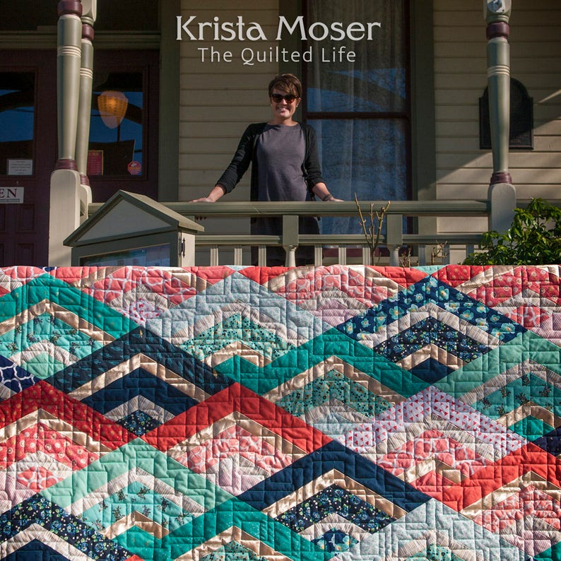 K&ouml;nnte beinhalten: Eine farbenfrohe Steppdecke mit einem Chevron-Muster in den Farben T&uuml;rkis, Blau, Rosa und Rot. Die Steppdecke ist aus verschiedenen Stoffen gefertigt und hat eine gesteppte Textur. Die Steppdecke wird auf einer Veranda ausgestellt, im Hintergrund steht eine Frau. Der Text "Krista Moser The Quilted Life" ist &uuml;ber der Frau zu sehen.