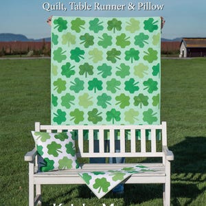May include: A light green quilt, table runner, and pillow set with a shamrock pattern. The quilt is held up behind a white bench, with a matching pillow and table runner on the bench. The text "CLOVER HILL" is at the top.