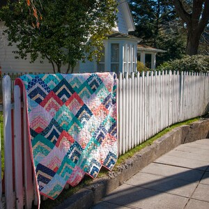 K&ouml;nnte beinhalten: Eine bunte Quiltdecke mit einem geometrischen Muster aus t&uuml;rkisfarbenen, rosafarbenen und goldenen Dreiecken h&auml;ngt an einem wei&szlig;en Lattenzaun. Die Quiltdecke ist aus Patchwork-Stoff gefertigt und hat einen gewellten Rand.