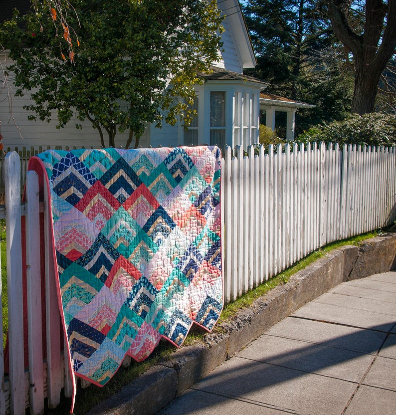 K&ouml;nnte beinhalten: Eine bunte Quiltdecke mit einem geometrischen Muster aus t&uuml;rkisfarbenen, rosafarbenen und goldenen Dreiecken h&auml;ngt an einem wei&szlig;en Lattenzaun. Die Quiltdecke ist aus Patchwork-Stoff gefertigt und hat einen gewellten Rand.