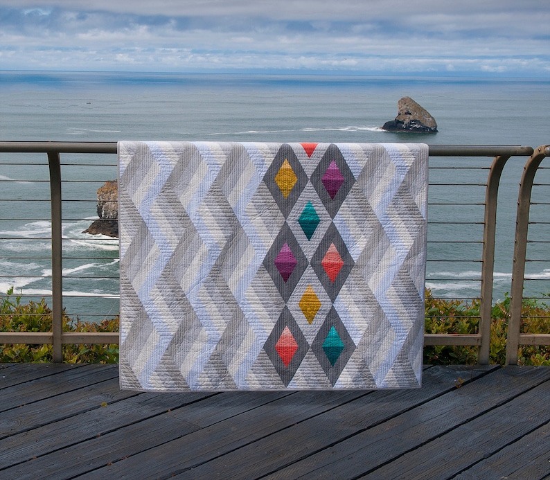 Puede incluir: Una colcha gris y blanca con un patr&oacute;n geom&eacute;trico de diamantes. Los diamantes est&aacute;n rellenos de tela colorida en tonos de amarillo, naranja, rojo, rosa, morado, azul verdoso y azul.