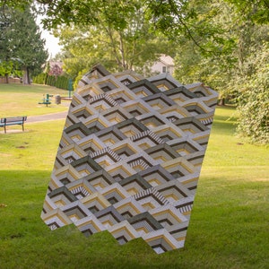 K&ouml;nnte beinhalten: Eine geometrisch gemusterte Steppdecke in Braun-, Wei&szlig;- und Gelbt&ouml;nen h&auml;ngt in einer grasbewachsenen Parklandschaft.