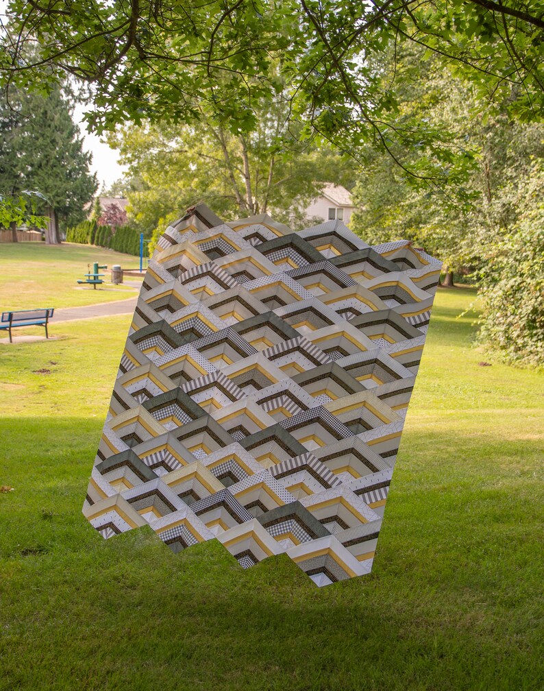 K&ouml;nnte beinhalten: Eine geometrisch gemusterte Steppdecke in Braun-, Wei&szlig;- und Gelbt&ouml;nen h&auml;ngt in einer grasbewachsenen Parklandschaft.