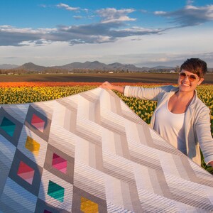 Puede incluir: Una mujer sostiene una gran colcha con un patr&oacute;n geom&eacute;trico de zigzags grises y blancos y diamantes de colores. La colcha est&aacute; delante de un campo de tulipanes amarillos y naranjas.