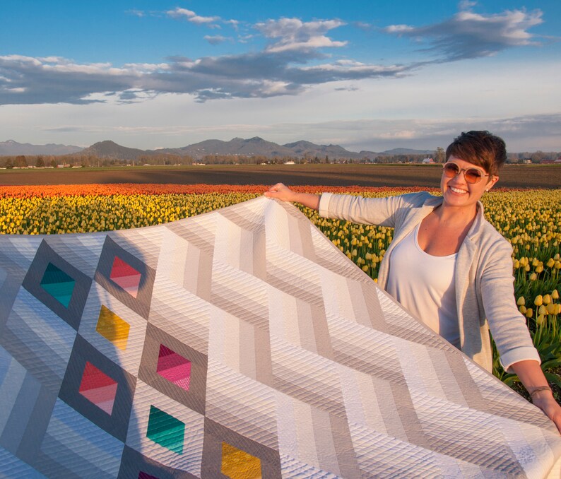 Puede incluir: Una mujer sostiene una gran colcha con un patr&oacute;n geom&eacute;trico de zigzags grises y blancos y diamantes de colores. La colcha est&aacute; delante de un campo de tulipanes amarillos y naranjas.