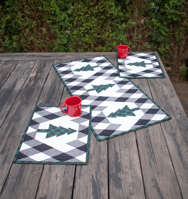 Puede incluir: Tres tazas rojas sobre un juego de tres manteles individuales acolchados verde y blanco con un patr&oacute;n geom&eacute;trico. Los manteles individuales presentan un dise&ntilde;o estilizado de &aacute;rbol de Navidad verde. Los manteles individuales est&aacute;n sobre una mesa de madera.