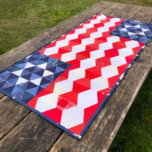 May include: A red, white, and blue quilted table runner with a chevron pattern on one side and a geometric pattern on the other side.