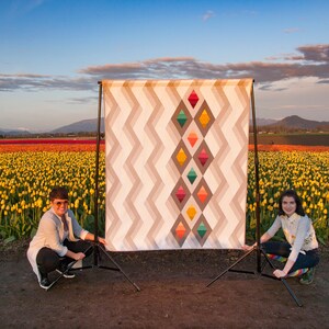 Puede incluir: Dos personas posan frente a una gran colcha con un patr&oacute;n geom&eacute;trico de diamantes en tonos de naranja, amarillo, verde y azul. La colcha se exhibe en un tr&iacute;pode en un campo de tulipanes amarillos.