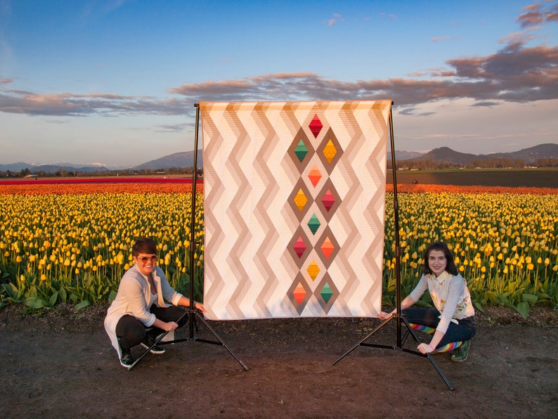 Puede incluir: Dos personas posan frente a una gran colcha con un patr&oacute;n geom&eacute;trico de diamantes en tonos de naranja, amarillo, verde y azul. La colcha se exhibe en un tr&iacute;pode en un campo de tulipanes amarillos.