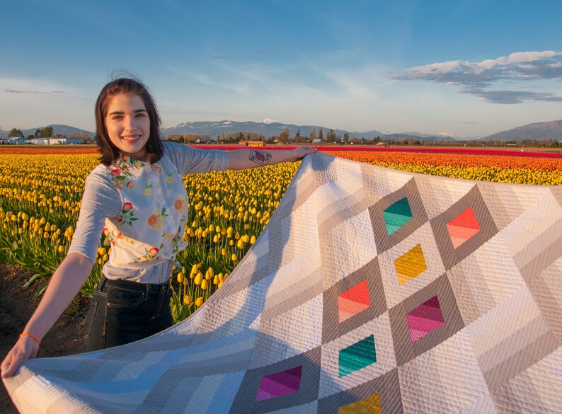 Puede incluir: Una persona sosteniendo una colcha grande con un patr&oacute;n geom&eacute;trico de diamantes grises, blancos y coloridos. La colcha est&aacute; en un campo de tulipanes amarillos.