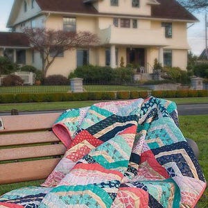 K&ouml;nnte beinhalten: Eine bunte Patchwork-Decke mit einem Chevron-Muster in Rosa-, Blau-, Gr&uuml;n- und Wei&szlig;t&ouml;nen liegt &uuml;ber einer Holzbank. Die Decke ist gefaltet und hat eine weiche, strukturierte Oberfl&auml;che.